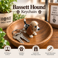 Bassett Hound keychain featuring a small 3D-printed dog figure in tan, white, and black, attached to a silver key ring with two keys, displayed on a wooden bowl with decorative background elements.