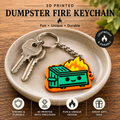 A 3D printed dumpster fire keychain displayed on a small beige ceramic dish over a wooden surface. The keychain features a green dumpster with orange flames rising from it, attached to a silver metal chain with two keys.