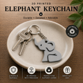 A 3D printed elephant keychain in matte gray is displayed on a small beige ceramic dish over a wooden surface. The keychain includes a silver metal key ring with two keys and a dangling elephant charm.