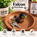 A 3D-printed bird-shaped keychain resting on a wooden dish, featuring a black bird with a yellow beak and brown body, attached to a silver keyring with two keys. The scene includes decorative props like a potted plant, a ceramic mug, and a framed sign, with text overlays reading 'Falcon Keychain' and feature icons at the bottom.