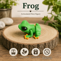 A green frog articulated flexi figure with orange feet sits on a round wood slice. The background is a styled tabletop scene with blurred green plants and stones, and text at the top reads 'Frog' and 'Articulated Flexi Figure.