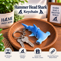 Blue hammer head shark keychain with attached metal key ring and keys, displayed on a wooden plate in a styled setting with decorative plants and a sign. Text at the top reads 'Hammer Head Shark Keychain' and feature icons appear along the bottom.