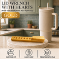 A gold metal lid wrench with a heart-shaped center is shown on a countertop next to a beige insulated tumbler. The background is a kitchen with green cabinets and blue-gray tile.