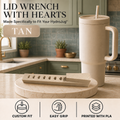 A tan insulated tumbler with a straw and handle stands on a round stone coaster in a kitchen setting, with a matching lid wrench tool placed in front. Text at the top reads 'LID WRENCH WITH HEARTS' and 'TAN,' with feature icons along the bottom.