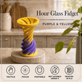 Hour glass fidget toy in purple and yellow, displayed on a marble coaster on a kitchen countertop with a blurred kitchen background. Text overlays read 'Hour Glass Fidget', 'PURPLE & YELLOW', and there is a feature icon row at the bottom.