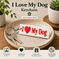 A white rectangular keychain with red text reading 'I [heart] My Dog' displayed on a beige ceramic dish with two keys attached. The scene includes decorative plants, a wooden surface, and marketing text at the top and bottom describing the product and care instructions.