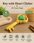 A yellow 3D-printed key-shaped fidget toy with a green heart in the center, displayed on a wooden surface with a bamboo steamer, chopsticks, and a cloth. The image includes text reading 'Key with Heart Clicker', '3D Printed Fidget', 'GREEN HEART', and feature icons for eco-friendly PLA, 3D printed PLA, fidget fun, great gift idea, and not suitable for children under 3.