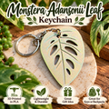 Monstera Adansonii leaf-shaped keychain with a silver metal key ring, displayed on a stone coaster with green leaves and a wooden surface. The image includes text reading 'Monstera Adansonii Leaf Keychain' and four feature icons at the bottom.