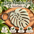 A large monstera leaf keychain made of metallic silver-toned material, displayed on a wood slice with decorative green leaves and text overlays reading 'Large Monstera Leaf Keychain' and feature icons below.
