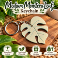 A monstera leaf-shaped keychain with a silver metal key ring and chain, displayed on a wooden block with decorative green leaves and text overlays reading 'Medium Monstera Leaf Keychain' and feature icons below.