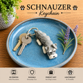 A 3D-printed gray Schauzer dog keychain resting on a blue ceramic plate with two keys, set on a wooden table with decorative plants and text overlays.