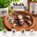 A sloth-shaped keychain displayed on a wooden tray, attached to metal keys. The keychain features a 3D sloth figure with brown and cream coloring and four curved claws.