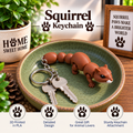 A brown 3D-printed squirrel keychain displayed on a green ceramic dish with two keys. The background features a potted plant and a framed sign, with text overlays highlighting the product's features.
