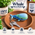A whale-shaped keychain in blue and green PLA plastic sits on a wooden tray with two keys. The scene includes decorative plants, a sign reading 'Whales Make a Brighter World,' and text overlays that say 'Whale Keychain' plus feature callouts: '3D Printed in PLA,' 'Detailed Design,' 'Great Gift for Ocean Lovers,' and 'Sturdy Keychain Attachment.